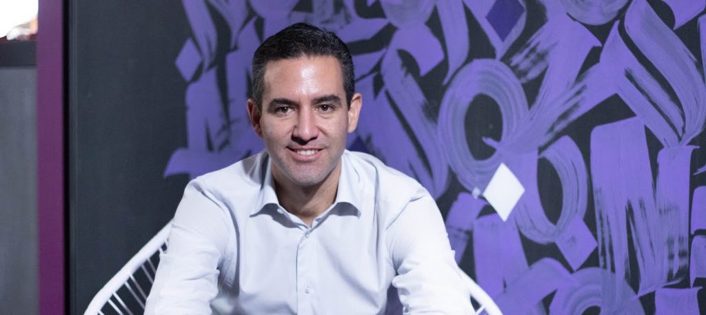 Portrait of David Vélez, founder and CEO of Nubank, sitting on a white chair with a purple and black background.